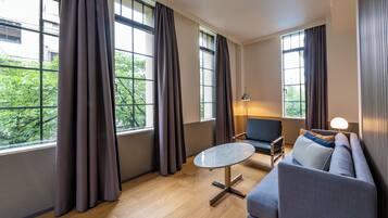 A modern living room with a sofa, a round table, and a lamp. Large windows with curtains are visible.