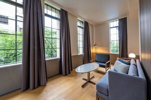 A modern living room with a sofa, a round table, and a lamp. Large windows with curtains are visible.