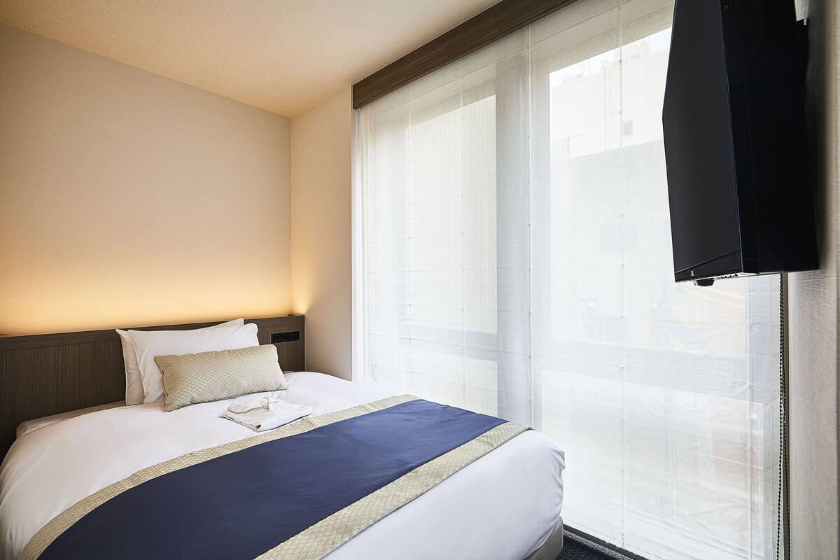 Una habitación de hotel con una cama bien hecha, un ventanal con persianas y un televisor montado en la pared.
