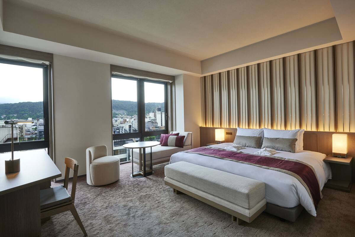 Una habitación de hotel moderna con una cama grande, un escritorio, una silla y vistas de la ciudad a través de amplias ventanas.