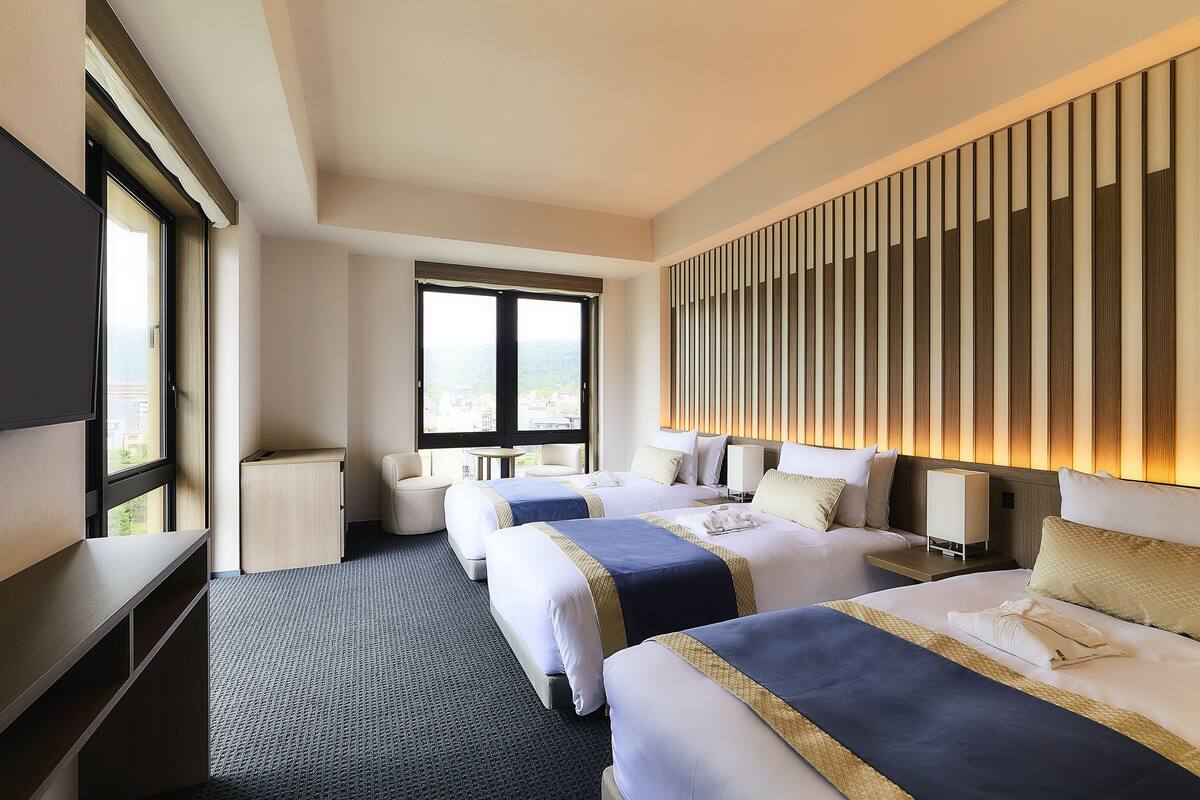 Habitación de hotel con dos camas, un escritorio, una silla y un amplio ventanal con vistas a la ciudad.
