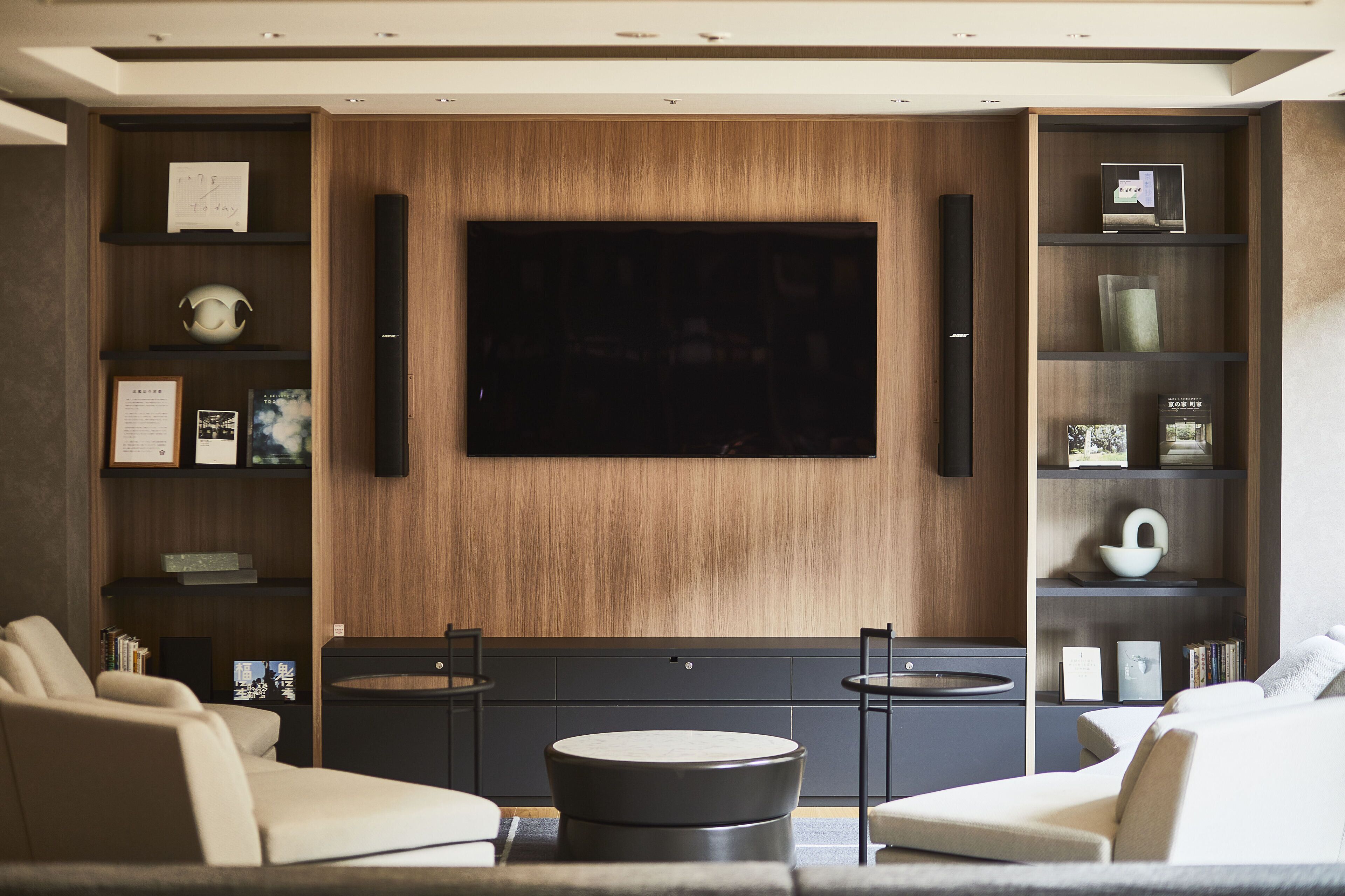 A modern living room with a large flat-screen TV mounted on a wooden wall, two armchairs, a round coffee table, and built-in shelving with decorative items.