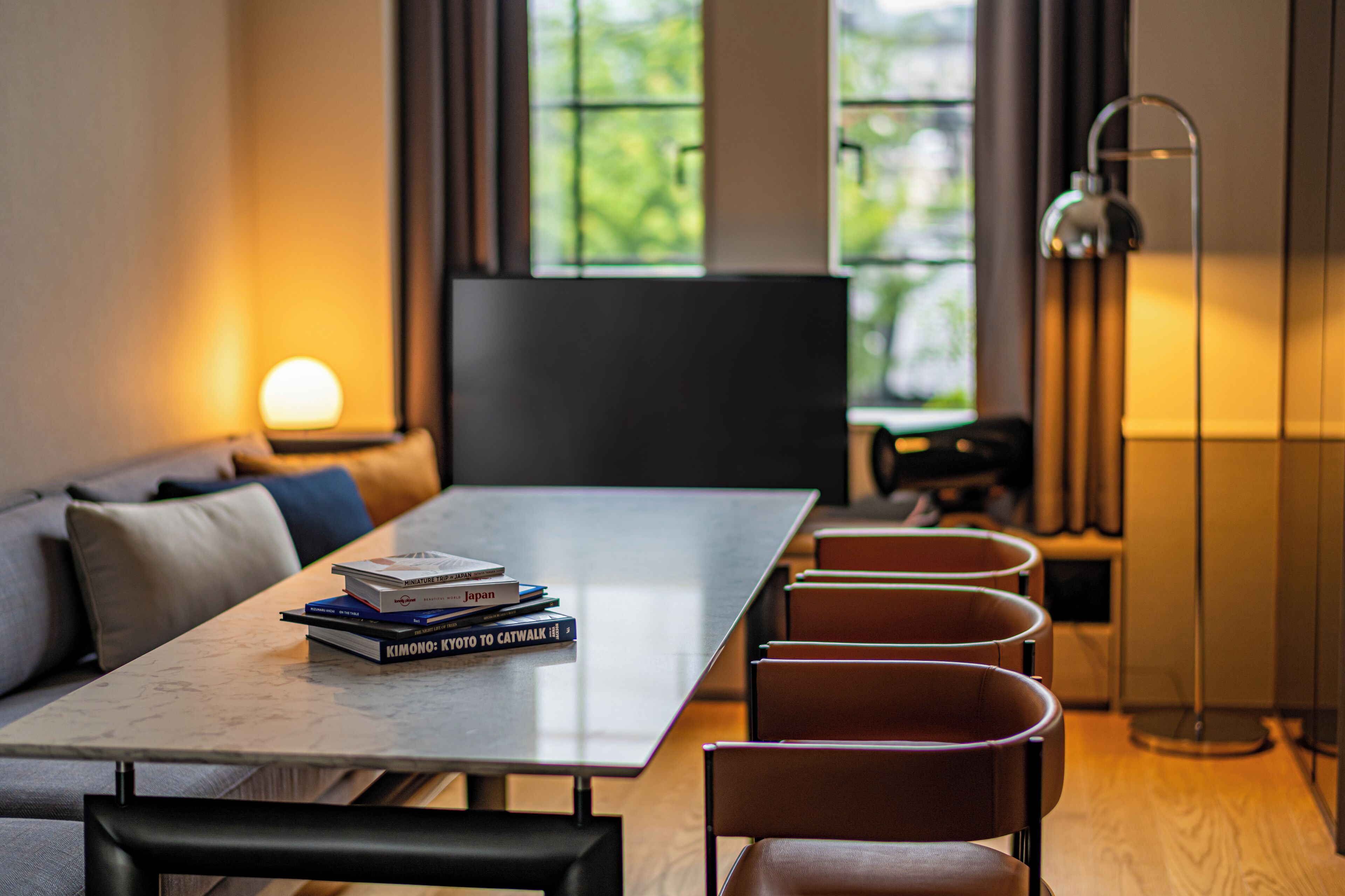 A modern room with a long table, a sofa, and a TV. There are books on the table and a lamp beside it.