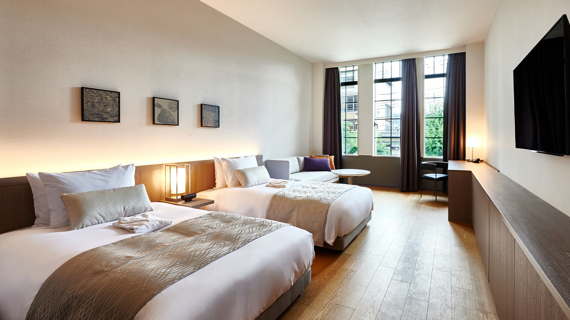 A hotel room with two beds, a desk, a chair, a TV, and large windows.