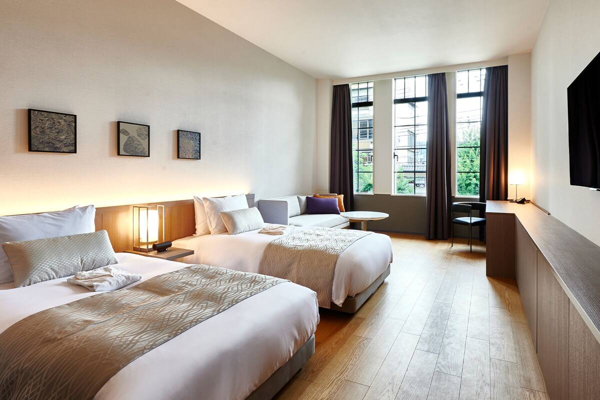 A hotel room with two beds, a desk, a chair, a TV, and large windows.
