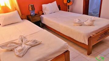 Dos camas individuales con ropa de cama y toallas blancas, una mesita de noche con lámpara y un ventilador de pared.