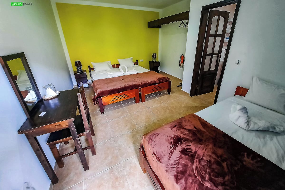 Quarto com duas camas, uma escrivaninha e um espelho. A parede é amarela e a porta tem um design triangular.