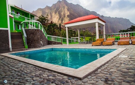 A swimming pool with a stone-tiled deck, a covered seating area with wooden loungers, and a building with a green facade and a red roof.
