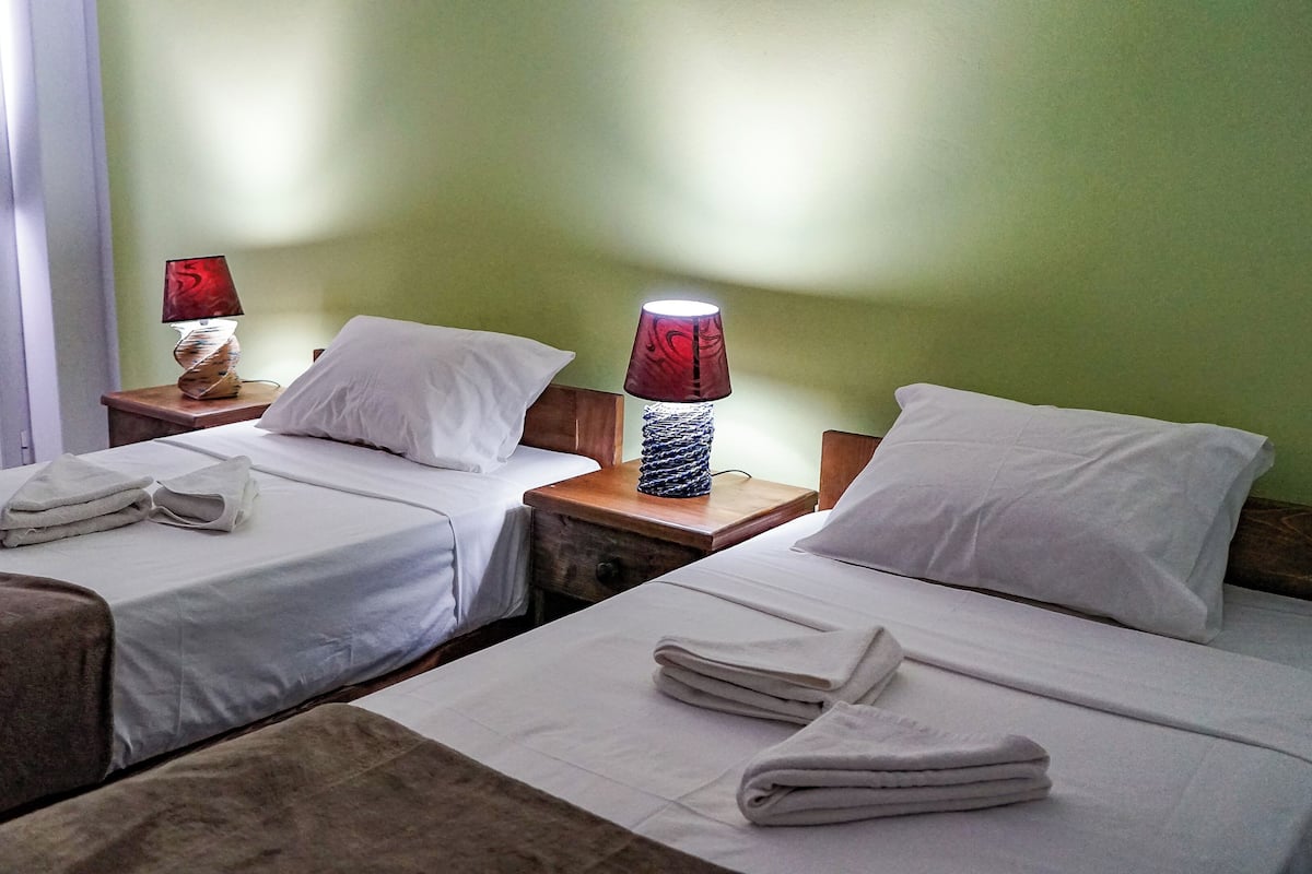 Quarto de hotel com duas camas de solteiro, cada uma com colcha e almofadas brancas, uma mesa de cabeceira de madeira com abajur vermelho e uma janela com cortinas brancas.