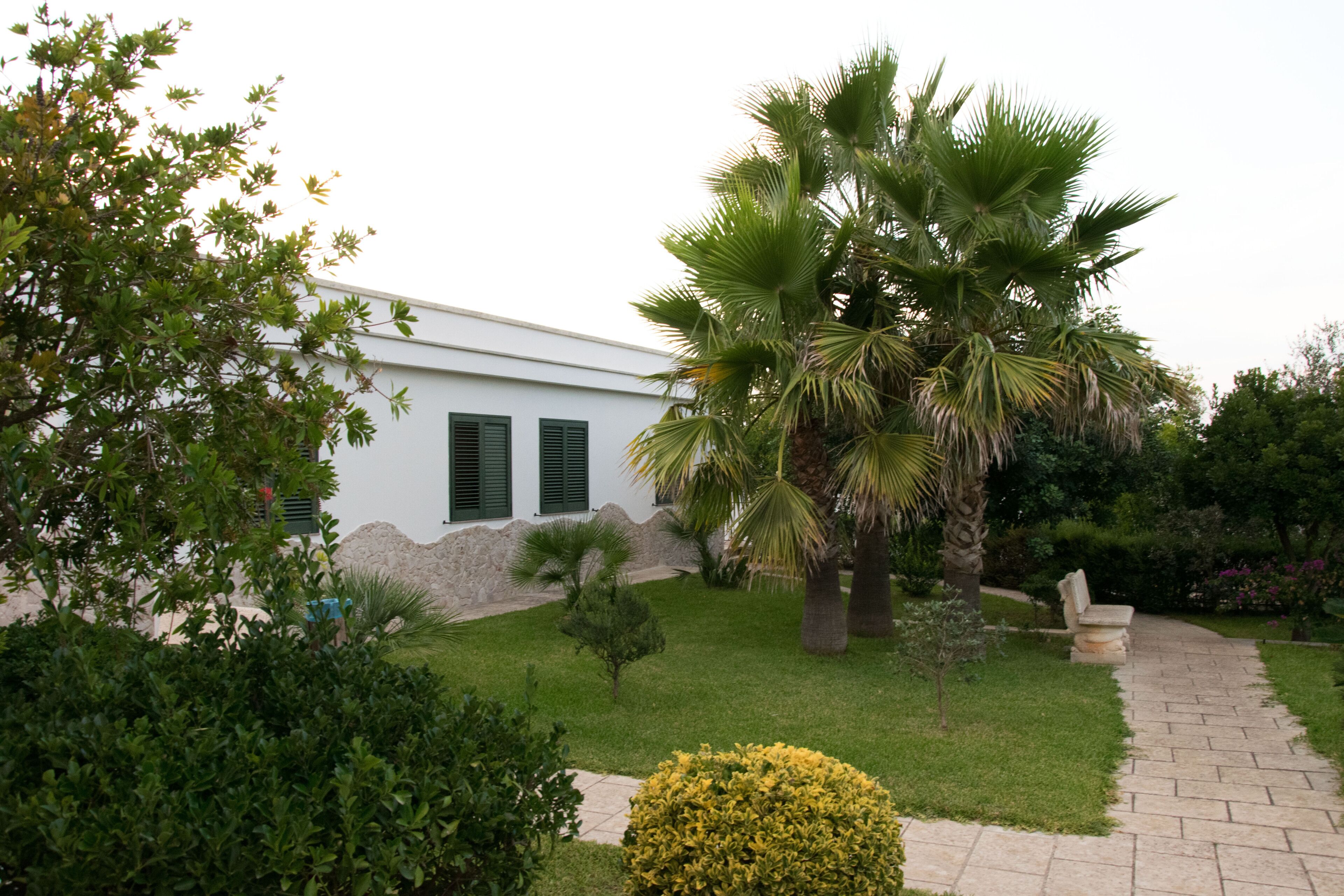 A white building with green shutters, surrounded by a well-maintained garden with palm trees, a stone wall, and a paved pathway.