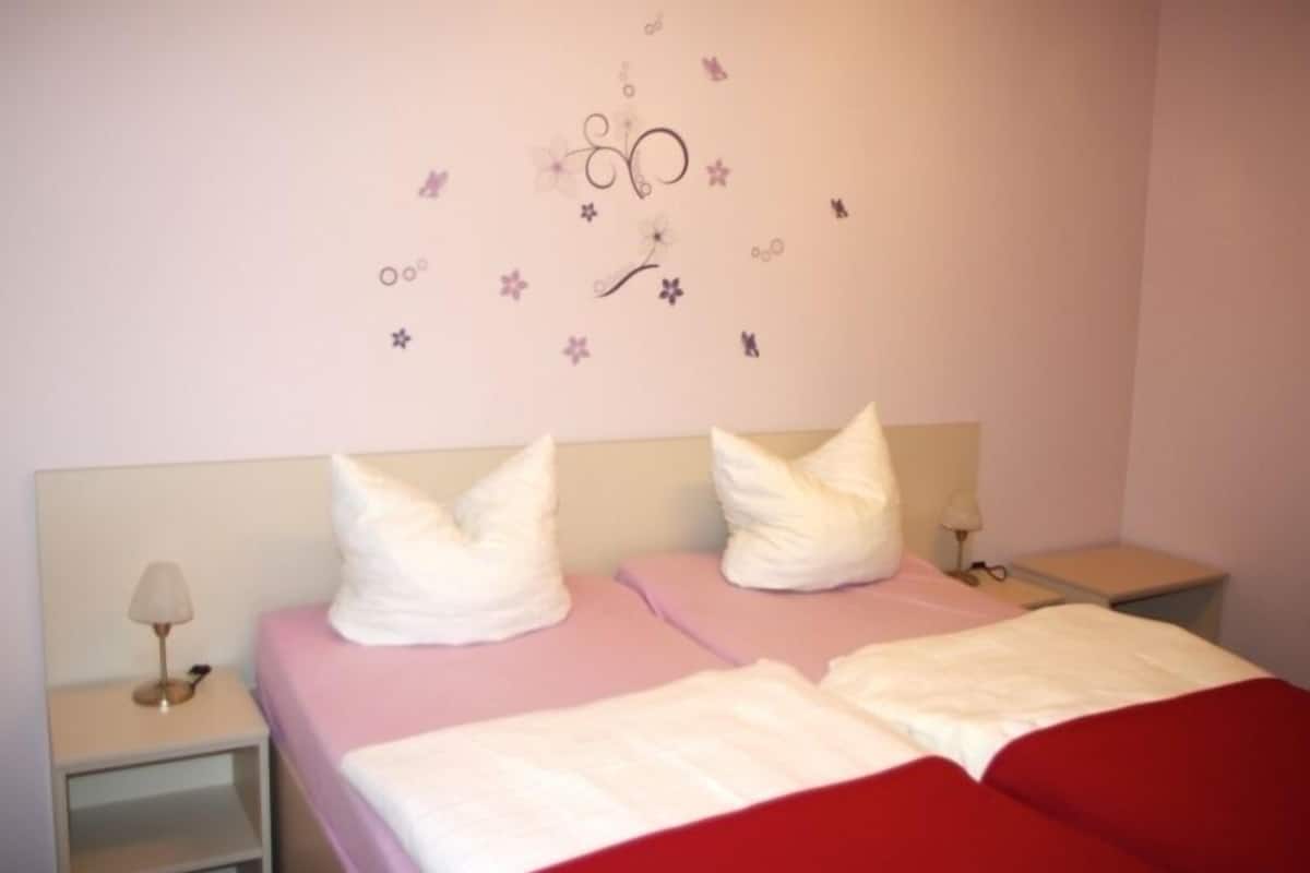 A bedroom with two beds, each with white pillows and pink bedding. There are bedside tables with lamps and a wall decoration above the beds.