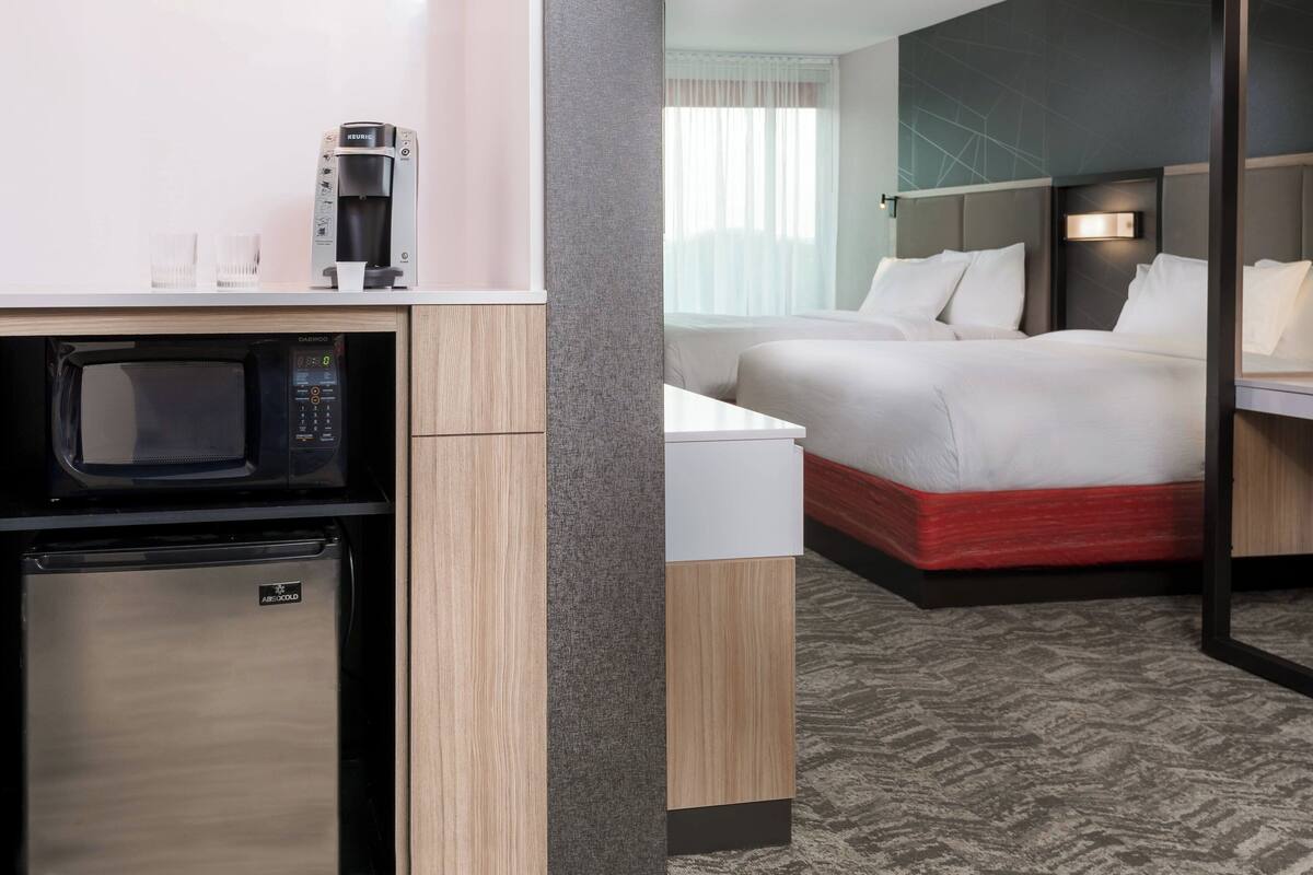 Een hotelkamer met een kitchenette, inclusief een magnetron, koelkast en koffiezetapparaat. Er staat een bed met wit beddengoed en een rode matras. De kamer heeft een modern design, met een gestructureerde grijze wand en een kamerbreed tapijt.