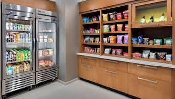 A kitchenette with two stainless steel refrigerators stocked with beverages and snacks, wooden cabinets with shelves holding various food items, and a countertop with a coffee machine.