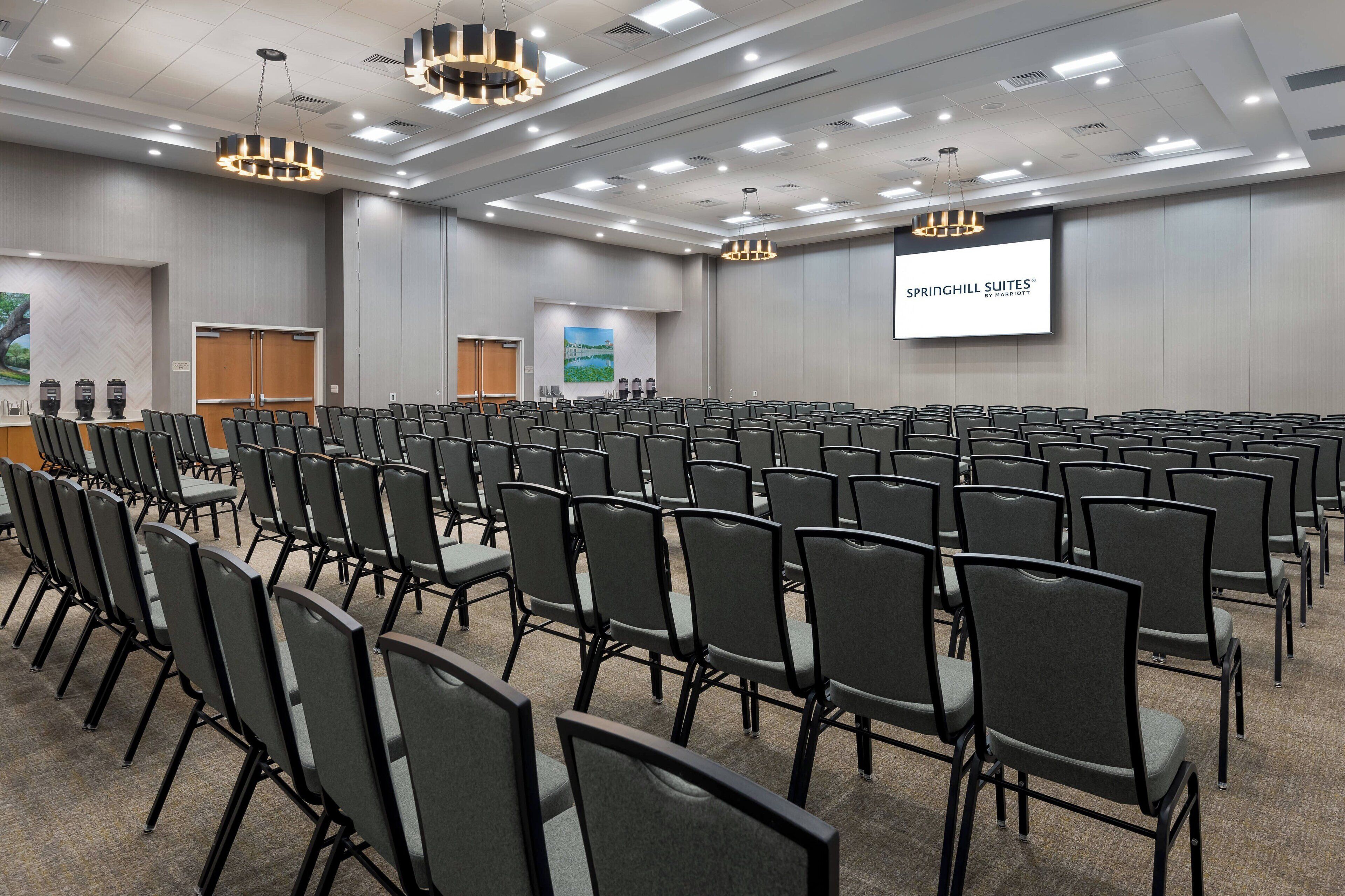 A conference room with rows of chairs, a projector screen displaying 'SPRINGHILL SUITES BY MARRIOTT', and a large screen with a landscape image.