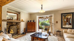 Un salon chaleureux avec une cheminée en pierre, un meuble en bois, un canapé confortable et une vue sur le jardin, visible à travers une porte vitrée.