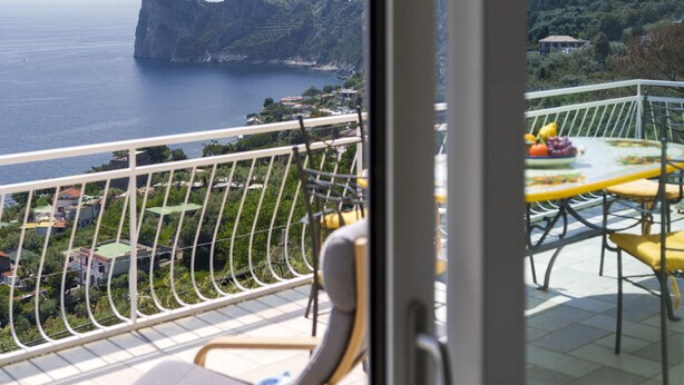 A balcony with a view of the sea and mountains, a table with chairs, and a hanging light fixture. - Belvedere Apartament in Nerano Stunning sea View (Massa Lubrense)