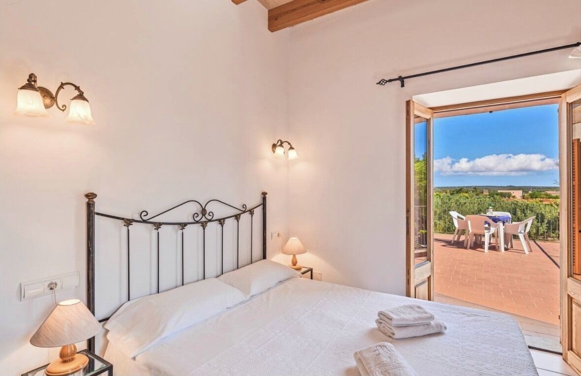 Quarto com estrutura de cama em metal, duas luminárias de cabeceira e vista para um pátio externo com mesa e cadeiras.
