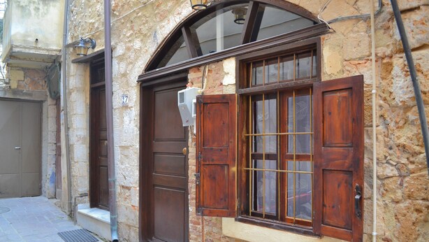 A narrow street with a building featuring a wooden door and window with shutters. - Yro Studios (Chania)
