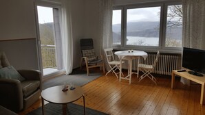 Una sala de estar acogedora con un sofá, una pequeña mesa redonda con vasos, un mueble para la televisión y vistas a un lago a través de amplios ventanales.