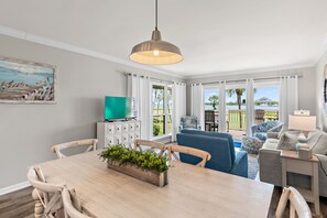A dining area with a wooden table, chairs, a plant centerpiece, a hanging light fixture, and a view of the outdoors through large windows.