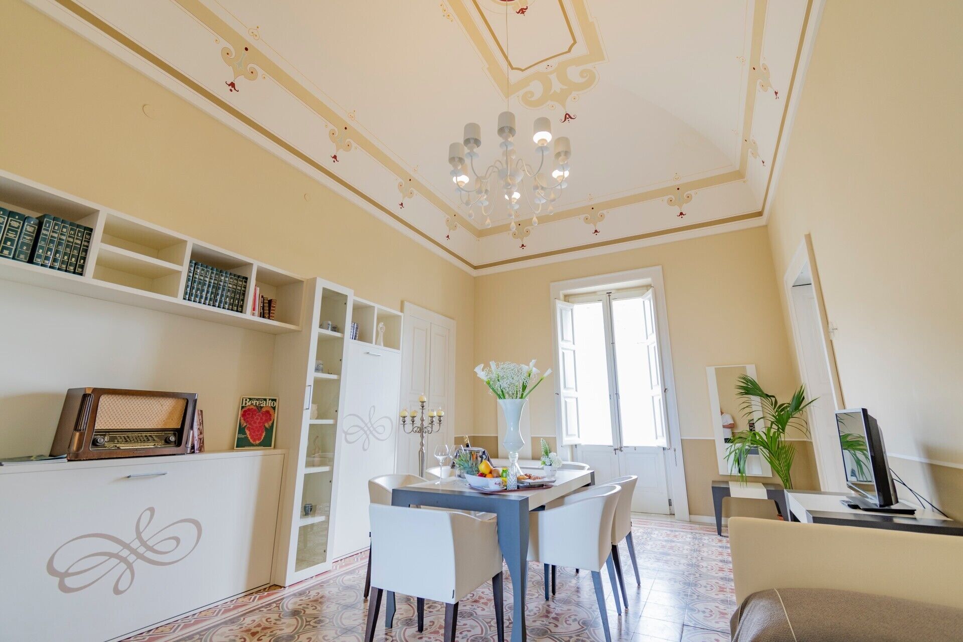 A dining area with a table set for two, a bookshelf, a chandelier, and a large window.