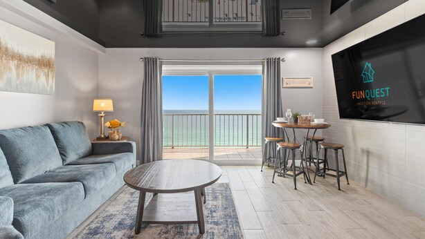 Uma sala de estar moderna com um sofá cinza, uma mesa de centro redonda e uma área de jantar com banquetas. Uma televisão grande exibe o logotipo "FUNQUEST Vacation Rentals".