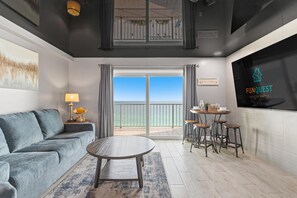 Un salon moderne avec un canapé gris, une table basse ronde et un coin repas équipé de tabourets. Un grand écran de télévision affiche le logo « FUNQUEST Vacation Rentals ».