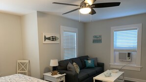 A bedroom with a bed, a dark gray sofa, a white nightstand, a white side table, a ceiling fan, and a window with blinds.