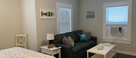 A bedroom with a bed, a dark gray sofa, a white nightstand, a white side table, a ceiling fan, and a window with blinds.