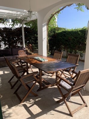 Eine Terrasse im Freien mit einem Holztisch und -stühlen, umgeben von weißen Wänden und mit einer Pergola bedeckt.