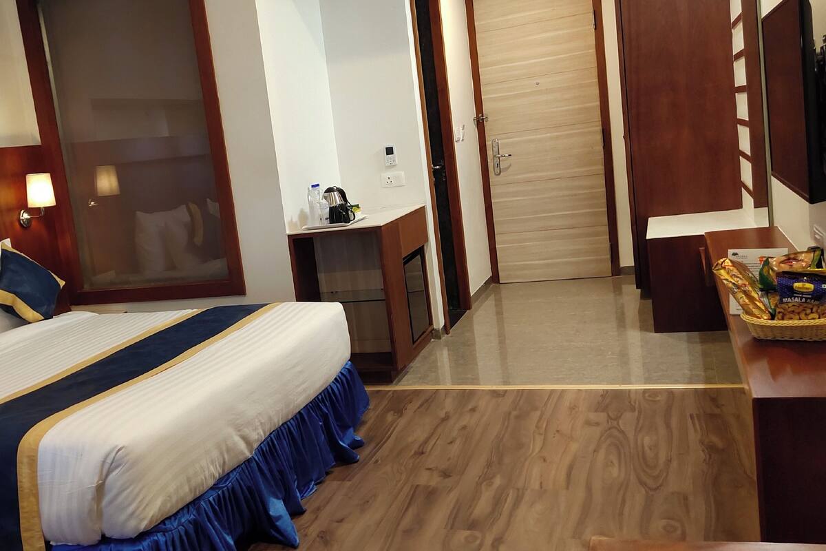 Une chambre d’hôtel avec un grand lit, une tête de lit en bois, une table de chevet avec une lampe, un petit bureau avec une chaise, une porte en bois et un téléviseur fixé au mur.