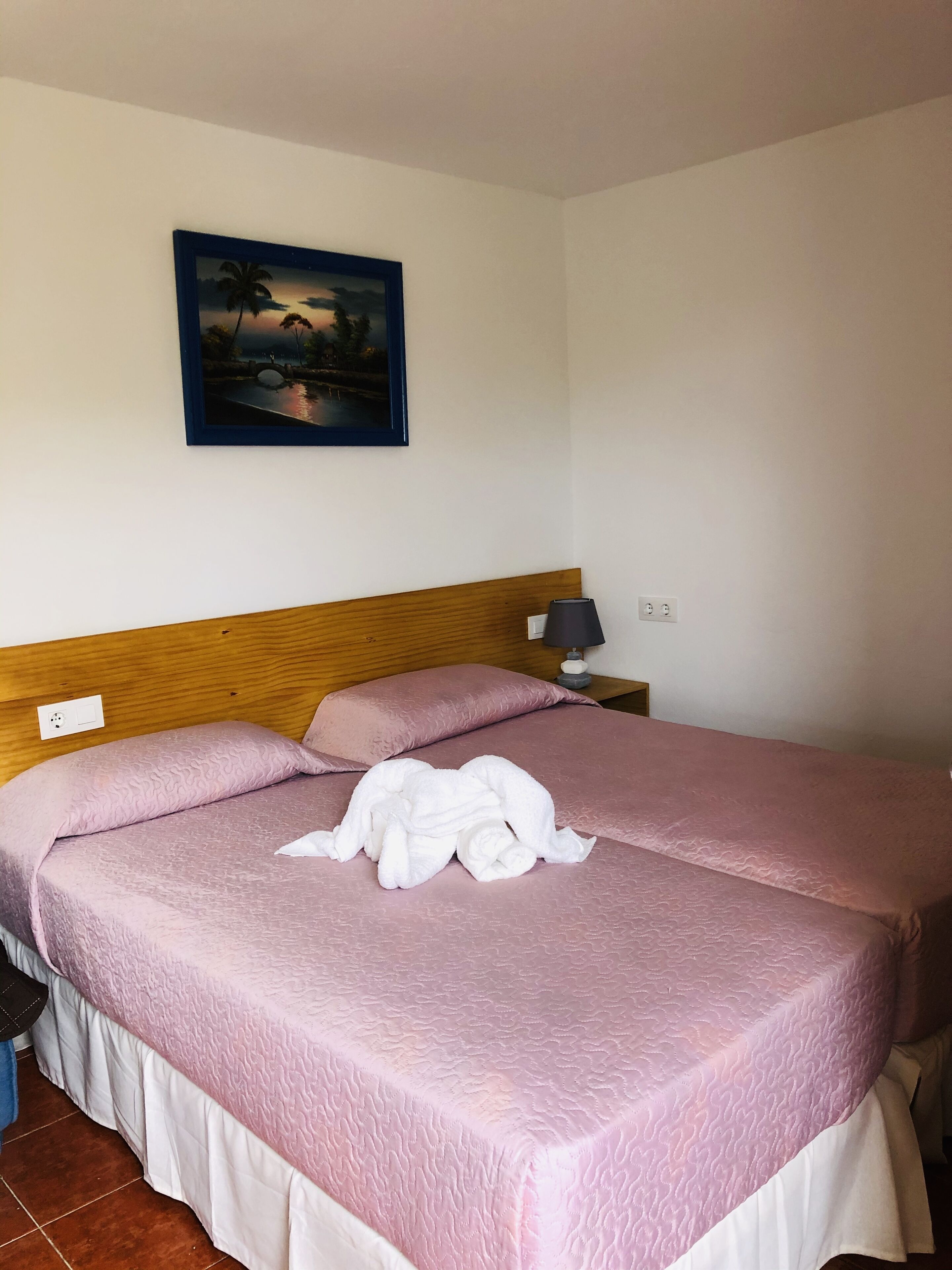 Ein ordentlich bezogenes Bett mit rosa Bettwäsche und einem weißen Handtuch darauf, ein hölzerner Kopfseitenwand, ein Nachttisch mit einer Lampe und ein Sonnenuntergangsbild an der Wand.