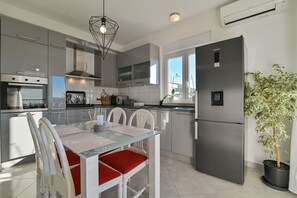 Eine moderne Küche mit grauen Schränken, einem Edelstahlbackofen, einem schwarzen Kühlschrank und einem Esstisch mit weißen Stühlen und roten Kissen.