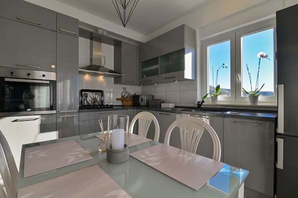 Eine moderne Küche mit Edelstahlgeräten, einem Essbereich mit Glastisch und Blick ins Freie durch große Fenster.