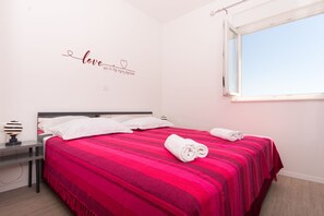 Ein ordentlich bezogenes Bett mit einer roten und rosafarben gestreiften Tagesdecke, weißen Kissen und einem aufgerollten weißen Handtuch daneben.