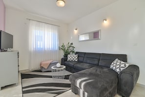 Ein modernes Wohnzimmer mit einem grauen Ecksofa, einem kleinen runden Couchtisch, einem Flachbildfernseher und einer Topfpflanze.
