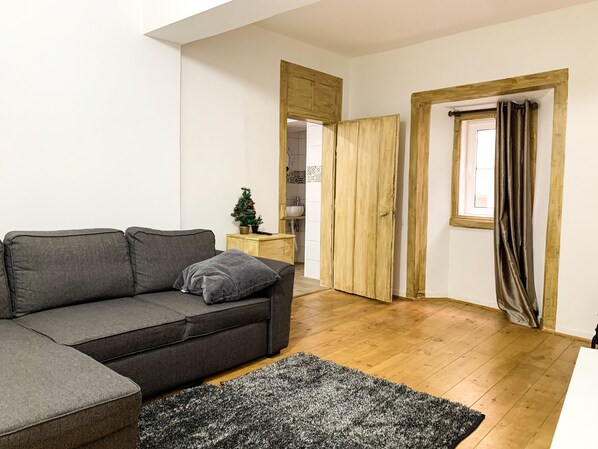 Una sala de estar con un sofá gris, una mesita con un árbol de Navidad, suelo de madera y una ventana con cortinas.