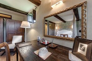 Una mesa de comedor de madera con un libro, una lámpara y un pequeño adorno decorativo. Un espejo con marco ornamentado refleja una pared de piedra y una cama con ropa blanca.