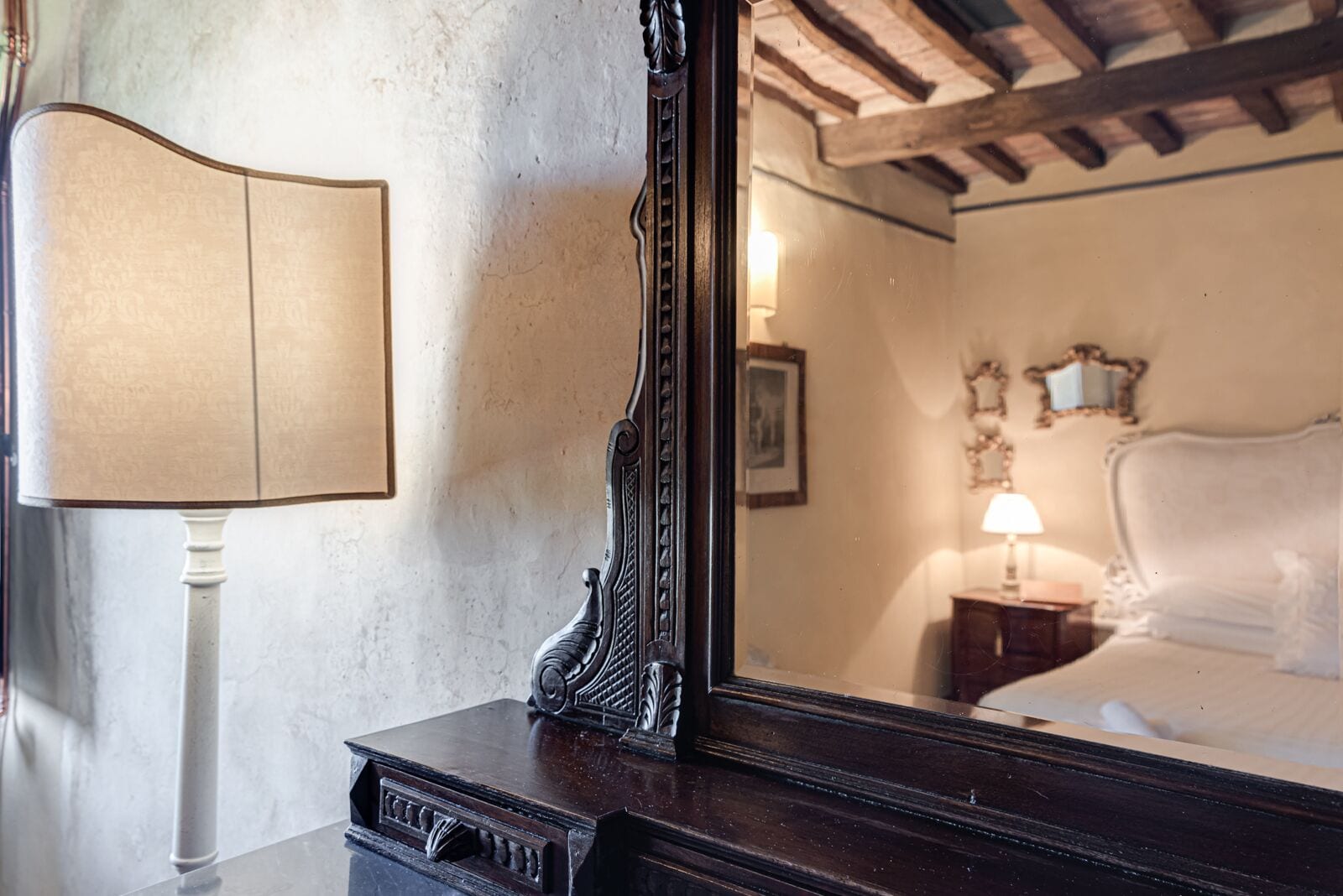 Un aparador de madera con un espejo que refleja un dormitorio. La habitación tiene una cama, una mesita de noche y una lámpara fijada a la pared.
