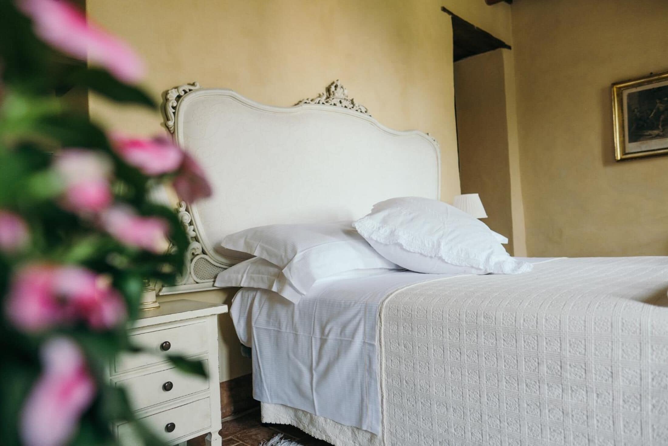 A bed with a white headboard and pillows, a nightstand with drawers, and a wall with a framed picture.