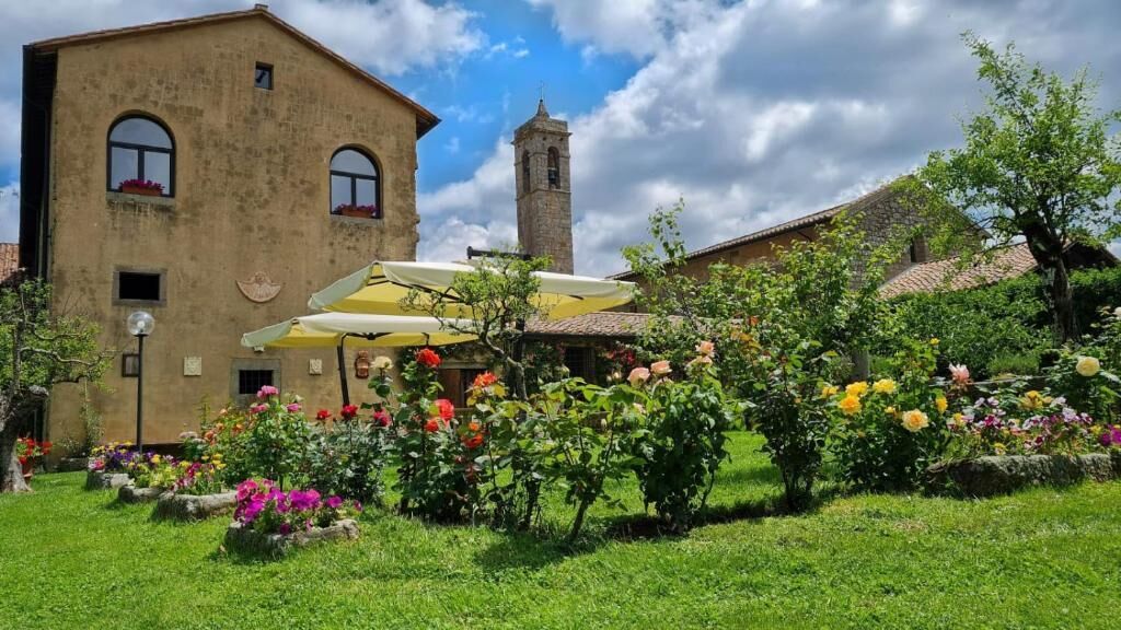 Ein Gebäude mit einem Glockenturm, umgeben von einem Garten mit verschiedenen Blumen und einem gelben Sonnenschirm.