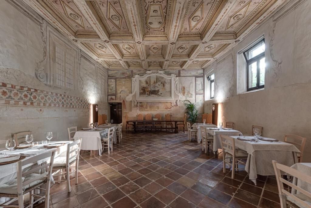 A spacious dining area with high ceilings and ornate ceiling decorations, featuring multiple tables and chairs arranged for dining.