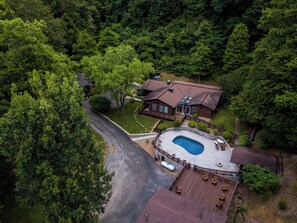 A residential property with a swimming pool, a covered deck area, and a house surrounded by dense forest. - Sprawling retreat-sleeps 27- private outdoor pool, hot tub, game room (Pigeon Forge)