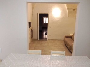 Ett rum med ett klinkergolv, ett vitt bord med två stolar, en beige soffa och en dörröppning som leder till ett annat rum.