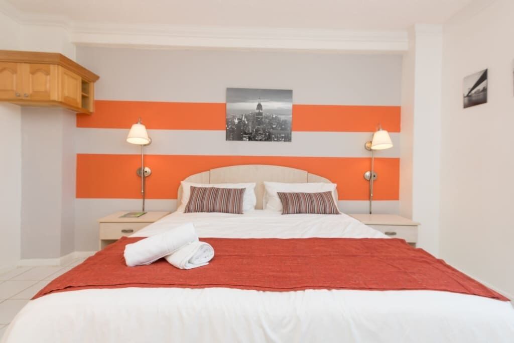 A hotel room with a bed, two bedside tables, a red throw blanket, and a cityscape picture on the wall.