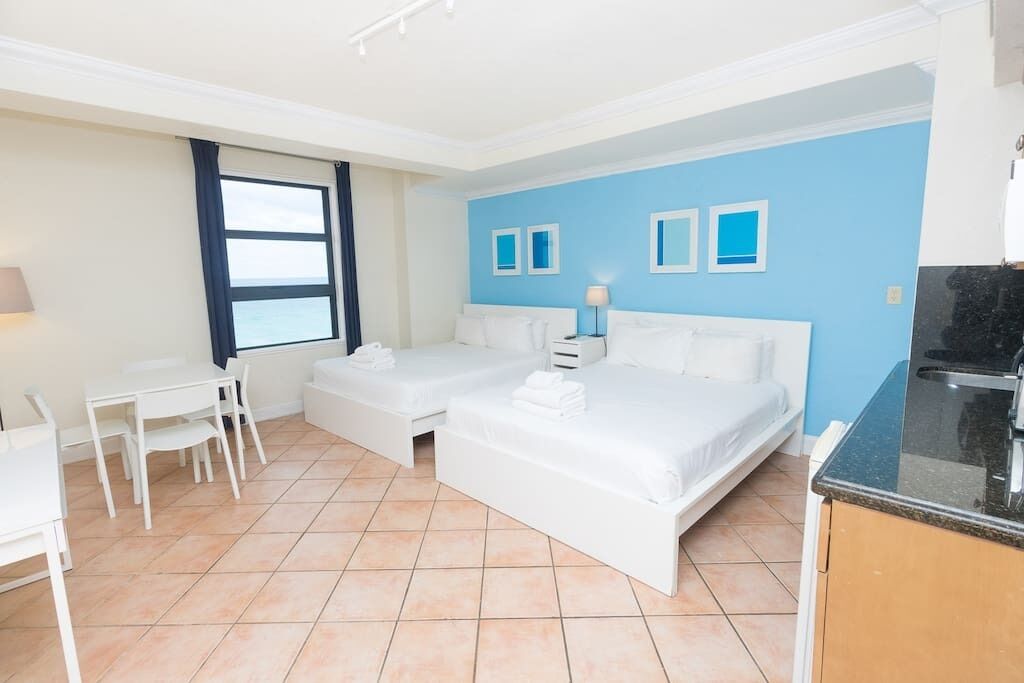 A hotel room with two beds, a desk, a chair, and a kitchenette.