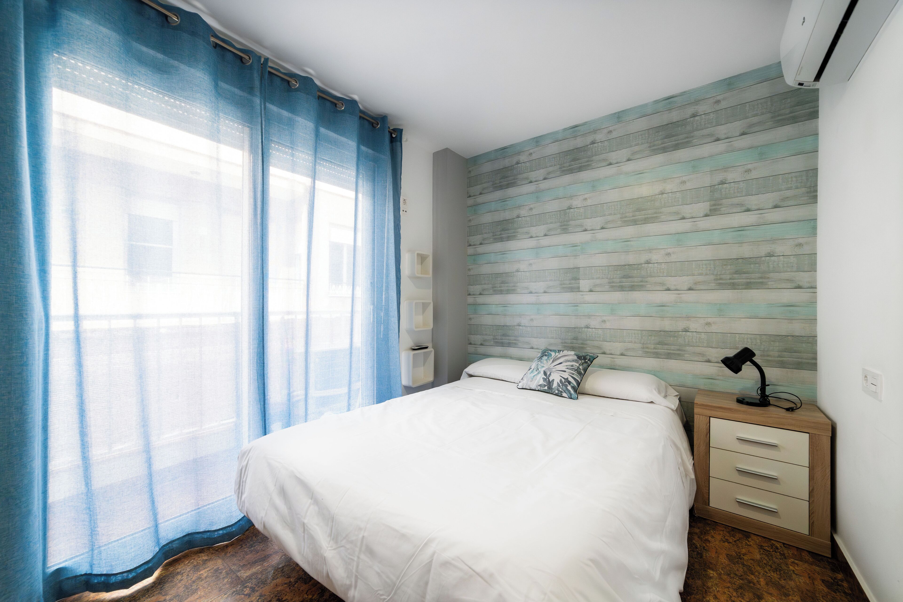 Un dormitorio con cama, mesita de noche, lámpara y ventana con cortinas.