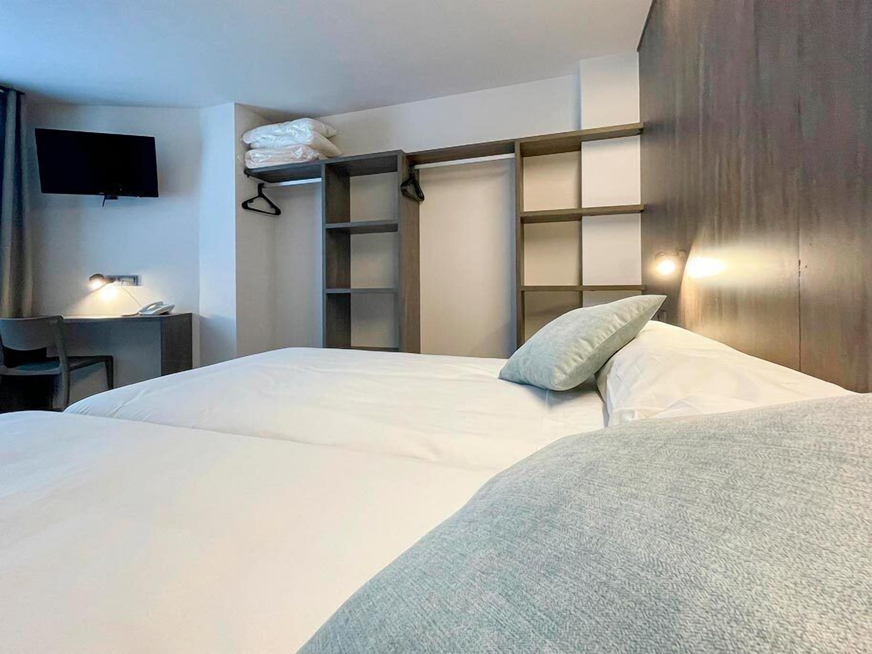 Ein Hotelzimmer mit einem Bett, einem Schreibtisch mit Lampe, einem an der Wand befestigten Fernseher und einem Kleiderschrank mit Regalen.