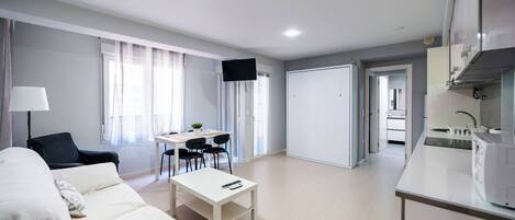Una sala moderna con un sofá, una mesa de comedor y un televisor. Hay una zona de cocina con un microondas y un fregadero.