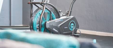 Una bicicleta estática con un ventilador grande sujeto en la parte trasera, una botella de agua y una toalla en el suelo.